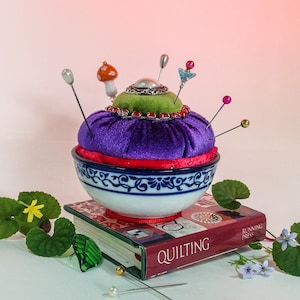 Könnte beinhalten: Ein violettes Samtkissen mit grünem Oberteil und rotem Besatz sitzt in einer blauen und weißen Schale. Die Schale steht auf einem Buch mit dem Titel "QUILTING" und dem Schriftzug "RUNNING PRESS" auf dem Rücken. Mehrere Nadeln und Stecknadeln stecken im Kissen.
