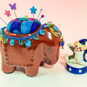 May include: A brown ceramic elephant pincushion with a blue and black fabric pincushion on top. The elephant is decorated with blue and green beads and a colourful jeweled pendant. A small porcelain clown figurine stands on a blue and white base with white stars.