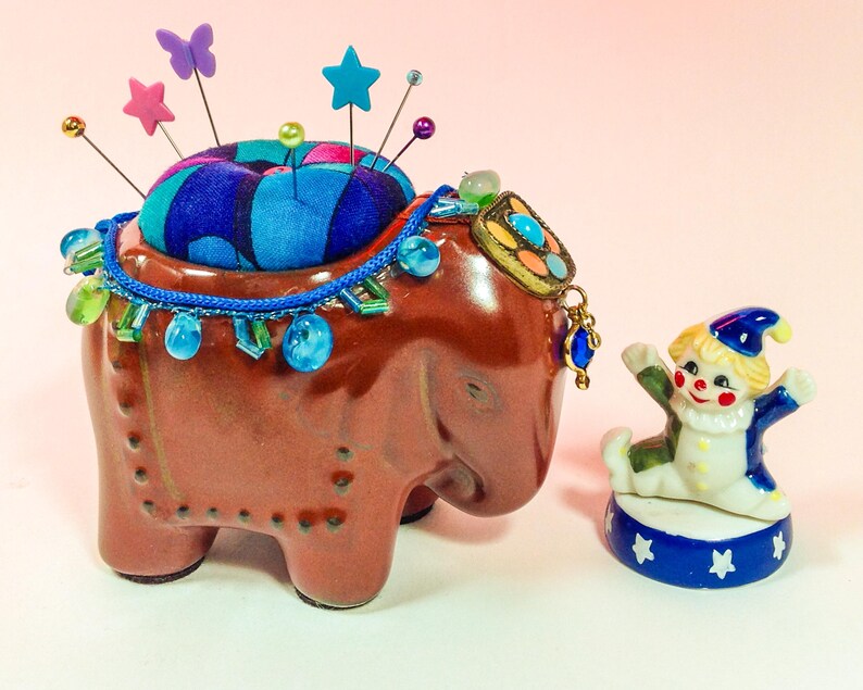 May include: A brown ceramic elephant pincushion with a blue and black fabric pincushion on top. The elephant is decorated with blue and green beads and a colourful jeweled pendant. A small porcelain clown figurine stands on a blue and white base with white stars.