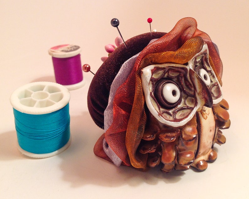 May include: A ceramic owl pincushion with brown, orange, and white details. The owl is wearing a brown velvet hat and a multi-colored ribbon. The pincushion is surrounded by two spools of thread, one purple and one blue.