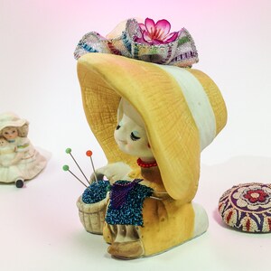 K&ouml;nnte beinhalten: Eine Keramikfigur eines M&auml;dchens, das einen gelben Hut mit einer rosa Blume tr&auml;gt und einen Korb mit blauen Perlen h&auml;lt. Die Figur ist ein Nadelkissen mit mehreren Nadeln, die aus dem Korb herausragen.