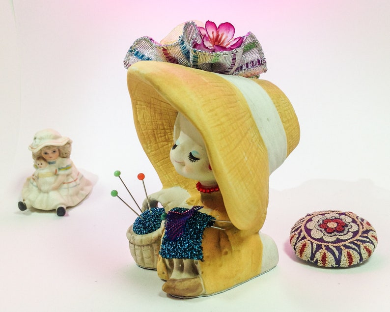 K&ouml;nnte beinhalten: Eine Keramikfigur eines M&auml;dchens, das einen gelben Hut mit einer rosa Blume tr&auml;gt und einen Korb mit blauen Perlen h&auml;lt. Die Figur ist ein Nadelkissen mit mehreren Nadeln, die aus dem Korb herausragen.