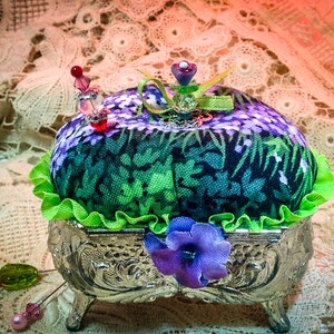 May include: A green and purple floral fabric pincushion with a green ruffle sits in a silver, ornate, three-legged stand. The pincushion is topped with a purple flower and a green ribbon.