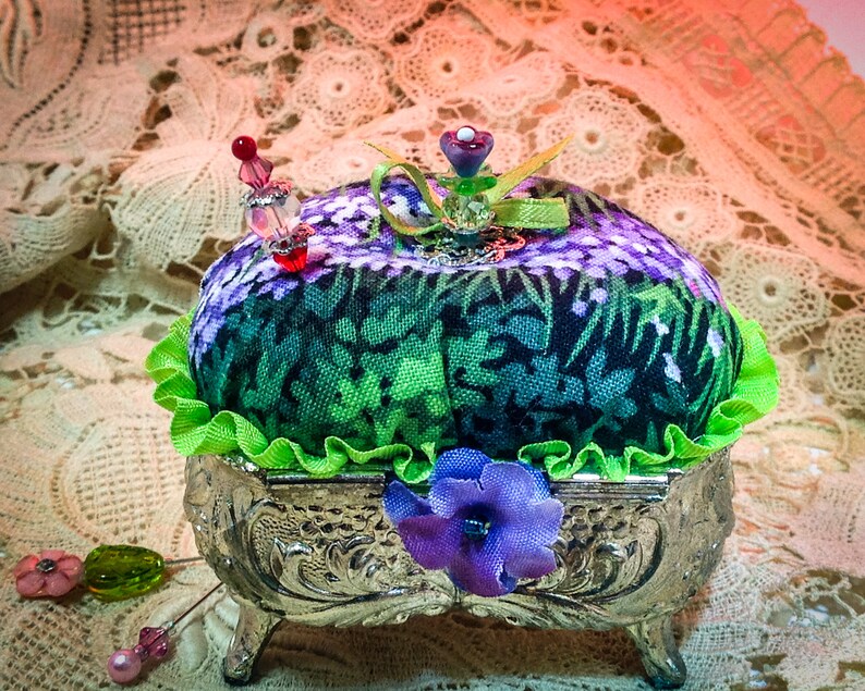 May include: A green and purple floral fabric pincushion with a green ruffle sits in a silver, ornate, three-legged stand. The pincushion is topped with a purple flower and a green ribbon.