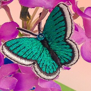 May include: A teal blue embroidered butterfly with white trim and a black center. The butterfly is sitting on a cluster of pink fabric flowers.