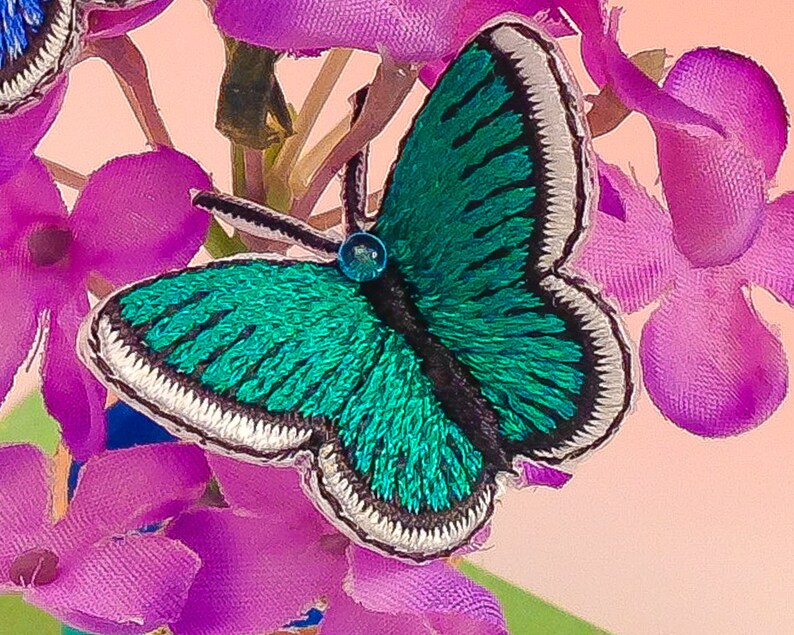 May include: A teal blue embroidered butterfly with white trim and a black center. The butterfly is sitting on a cluster of pink fabric flowers.
