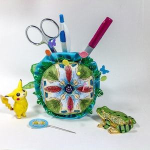 Può includere: Un cuscino per spilli colorato con un motivo floreale, circondato da una base in tessuto blu. Un paio di forbici, una penna e una matita sono infilate nel cuscino per spilli. Nell'immagine sono presenti anche una piccola statuina di Pikachu gialla e una statuina di rana verde.