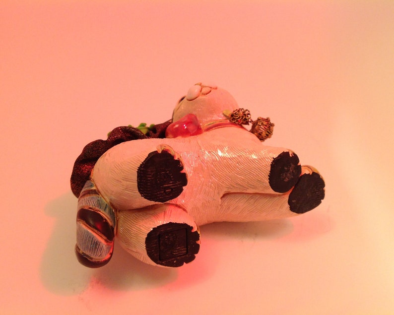 May include: A white ceramic figurine of a pig wearing a red and gold hat and a blue and gold scarf. The pig has black hooves and is lying on its side.