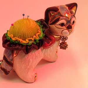 May include: A white ceramic cat figurine with a yellow and green pincushion on its back. The cat has a black and blue patterned face and a gold charm hanging from its neck.
