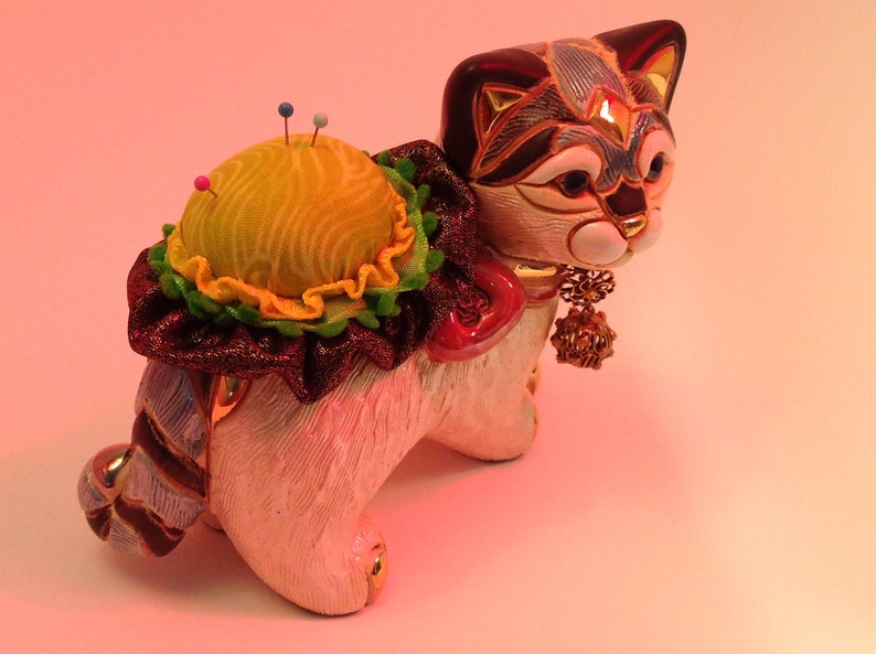 May include: A white ceramic cat figurine with a yellow and green pincushion on its back. The cat has a black and blue patterned face and a gold charm hanging from its neck.