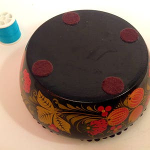 May include: A black wooden bowl with a floral design painted in red, yellow, and gold. The bowl has four felt pads on the bottom.