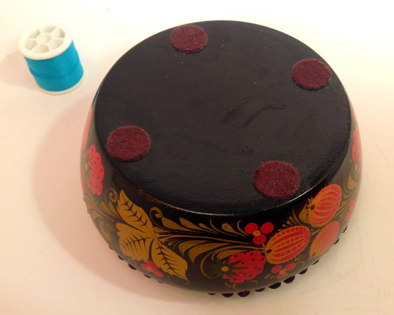 May include: A black wooden bowl with a floral design painted in red, yellow, and gold. The bowl has four felt pads on the bottom.