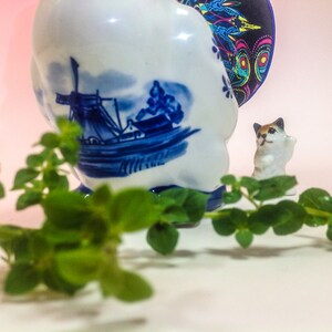 May include: A blue and white ceramic figurine of a cat with a windmill and boat design. The figurine is sitting on a white surface with green leaves in the foreground.