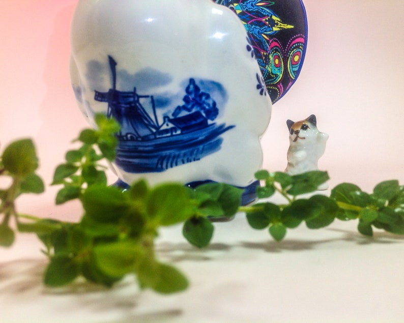 May include: A blue and white ceramic figurine of a cat with a windmill and boat design. The figurine is sitting on a white surface with green leaves in the foreground.