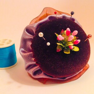 May include: A purple velvet pincushion with a flower design and multiple pins. The pincushion is surrounded by a light purple fabric with a brown trim.