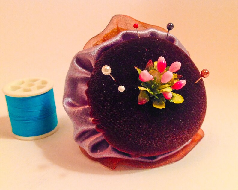 May include: A purple velvet pincushion with a flower design and multiple pins. The pincushion is surrounded by a light purple fabric with a brown trim.
