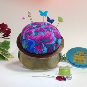 May include: A round pincushion with a floral fabric cover in shades of pink, blue, and green. The pincushion is sitting on a brass base and has several pins with colorful butterfly and leaf-shaped heads. The pincushion is sitting on a white surface with a spool of green thread and a sticker that says "PincushionPalace.com".