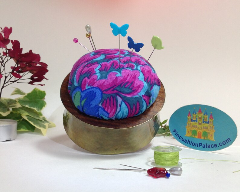 May include: A round pincushion with a floral fabric cover in shades of pink, blue, and green. The pincushion is sitting on a brass base and has several pins with colorful butterfly and leaf-shaped heads. The pincushion is sitting on a white surface with a spool of green thread and a sticker that says "PincushionPalace.com".