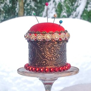 May include: A red pincushion with a gold and silver decorative band sits on a glass pedestal. The pincushion is surrounded by red beads.