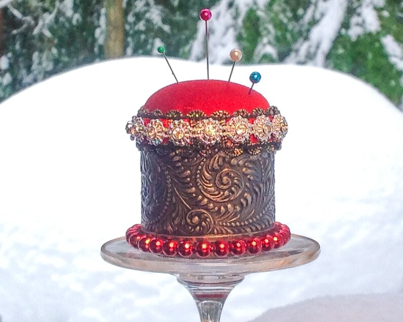 May include: A red pincushion with a gold and silver decorative band sits on a glass pedestal. The pincushion is surrounded by red beads.