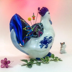 May include: A white ceramic rooster pincushion with blue and white floral designs. The rooster has a purple comb and a blue satin bow on its tail. The pincushion is filled with colourful pins and beads.