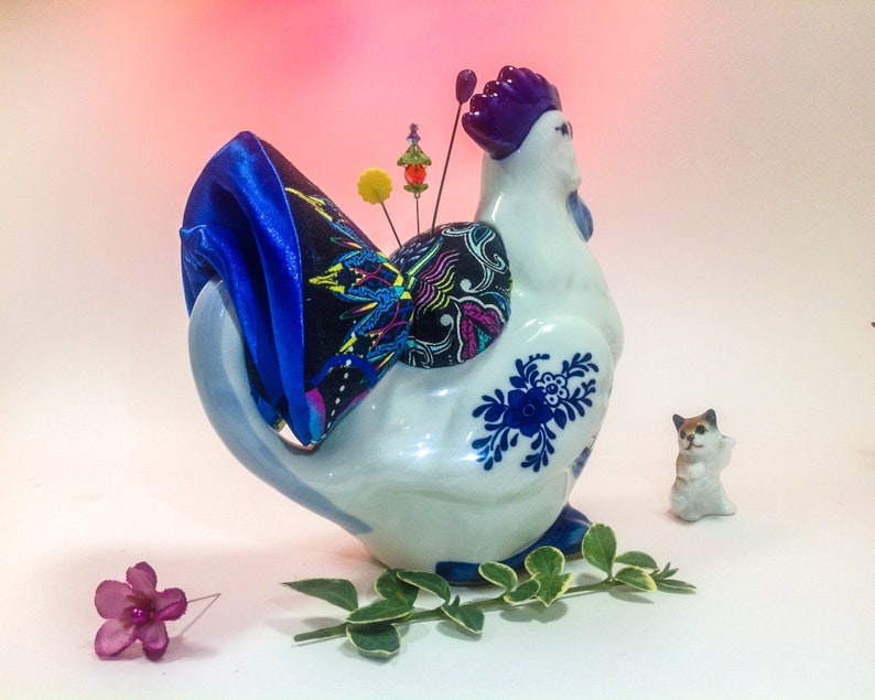 May include: A white ceramic rooster pincushion with blue and white floral designs. The rooster has a purple comb and a blue satin bow on its tail. The pincushion is filled with colourful pins and beads.