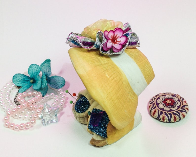 K&ouml;nnte beinhalten: Eine Keramikfigur einer Frau, die einen gelben Hut mit einer rosa Blume und einem wei&szlig;en Band tr&auml;gt. Die Figur h&auml;lt ein Nadelkissen mit bunten Nadeln und einer blau-wei&szlig;en Blume.