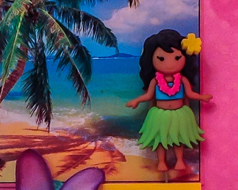 May include: A colorful figurine of a hula dancer with a green grass skirt, a blue top, and a pink flower lei. She has brown hair and a yellow flower in her hair. The background is a beach scene with a palm tree and blue water.