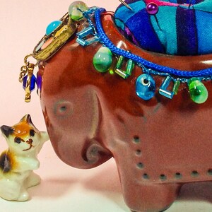 May include: A brown ceramic elephant pincushion with a blue and green beaded trim and a small white cat figurine. The elephant has a blue and green beaded trim around the neck and a small white cat figurine is standing next to it.