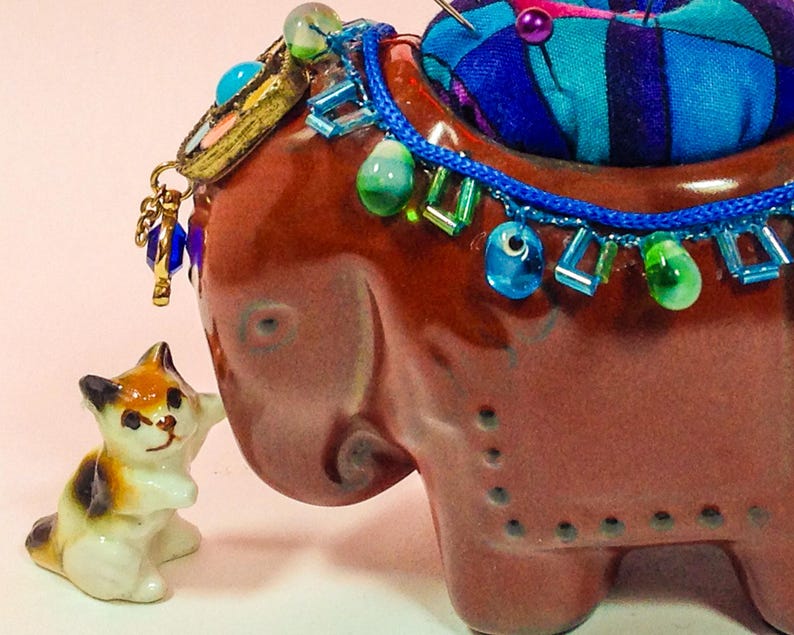 May include: A brown ceramic elephant pincushion with a blue and green beaded trim and a small white cat figurine. The elephant has a blue and green beaded trim around the neck and a small white cat figurine is standing next to it.