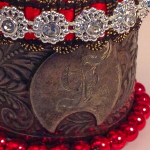 May include: A silver cuff bracelet with an ornate design and a large letter "F" in the center. The bracelet is decorated with red beads and a band of red ribbon with silver rhinestones.