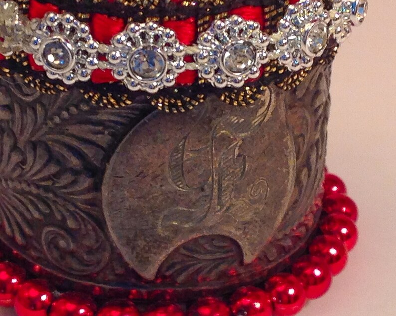 May include: A silver cuff bracelet with an ornate design and a large letter "F" in the center. The bracelet is decorated with red beads and a band of red ribbon with silver rhinestones.