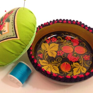 May include: A green pincushion with a floral design and a black and gold painted wooden bowl with a red and yellow floral design. The bowl has a black and red trim around the edge.