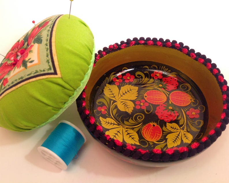 May include: A green pincushion with a floral design and a black and gold painted wooden bowl with a red and yellow floral design. The bowl has a black and red trim around the edge.