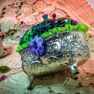 May include: A silver pincushion with a green and floral fabric top. The pincushion has three legs and is decorated with floral designs. There are several pins stuck in the top of the pincushion.