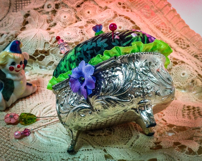 May include: A silver pincushion with a green and floral fabric top. The pincushion has three legs and is decorated with floral designs. There are several pins stuck in the top of the pincushion.