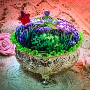 May include: A green, purple, and white floral pincushion with a green ruffle sits on a silver stand with three legs. The pincushion has a red star and a blue flower on top.