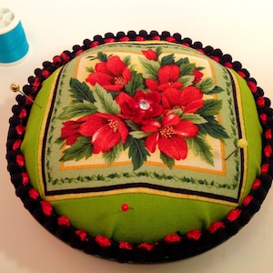 May include: A round pincushion with a green fabric top featuring a floral design with red flowers and green leaves. The pincushion is surrounded by a black and red trim.