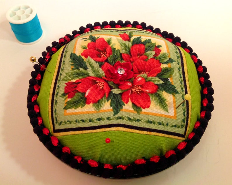 May include: A round pincushion with a green fabric top featuring a floral design with red flowers and green leaves. The pincushion is surrounded by a black and red trim.