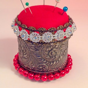May include: A red pincushion with a gold and silver trim sits on a base of red beads. The pincushion is decorated with rhinestones and has several pins sticking out of it.