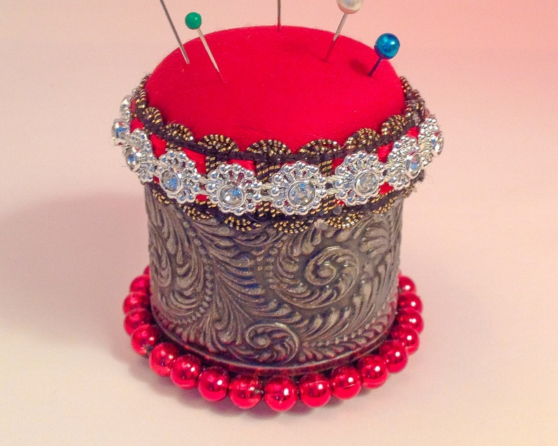 May include: A red pincushion with a gold and silver trim sits on a base of red beads. The pincushion is decorated with rhinestones and has several pins sticking out of it.