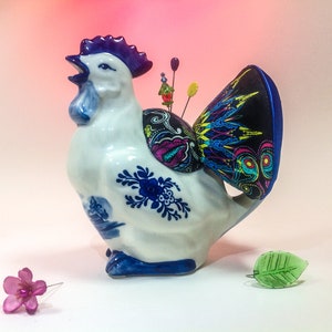 May include: A white ceramic rooster figurine with blue and white painted details. The rooster has a black and multi-coloured fabric pincushion on its back.