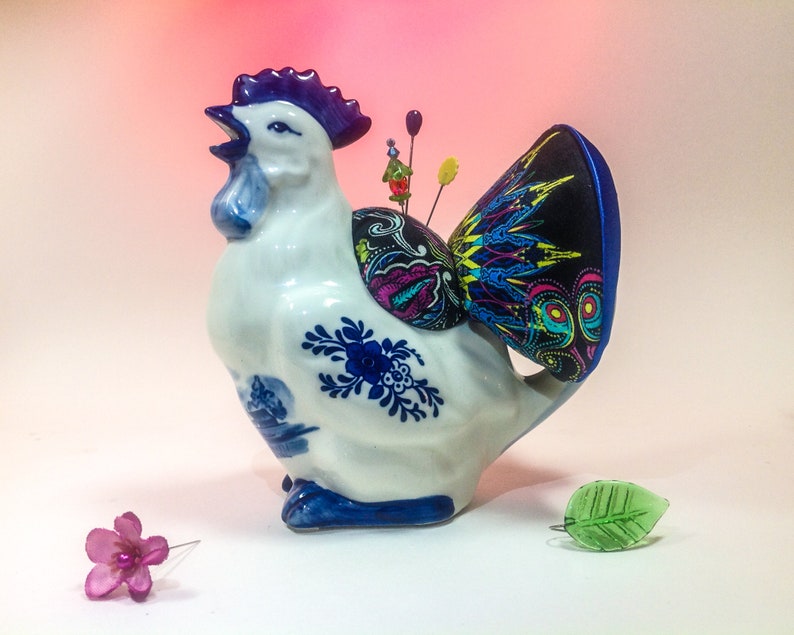 May include: A white ceramic rooster figurine with blue and white painted details. The rooster has a black and multi-coloured fabric pincushion on its back.