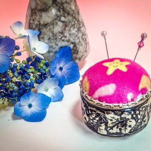 Op de afbeelding: Een decoratief speldenkussen met een felroze bovenkant met een lachend sterrenontwerp. Het speldenkussen staat op een zilverkleurige basis met een bladerenpatroon. Blauwe hortensia's en een marmeren vaas staan op de achtergrond.