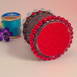 May include: A small, decorative, silver-colored container with a red, glittery bottom. The container is decorated with a beaded trim and rhinestones.