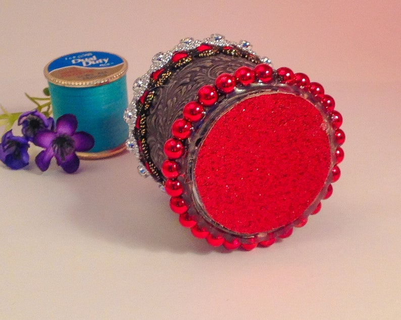 May include: A small, decorative, silver-colored container with a red, glittery bottom. The container is decorated with a beaded trim and rhinestones.