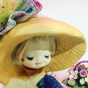 K&ouml;nnte beinhalten: Eine Porzellanpuppe mit einem gelben Hut und einem Strau&szlig; k&uuml;nstlicher Blumen. Die Puppe hat langes blondes Haar, blaue Augen und ein rot-wei&szlig;es Kleid.