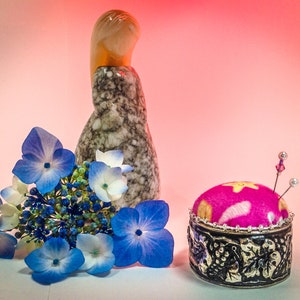 Op de afbeelding: Een stilleven met een roze en zilveren speldenkussen met spelden, een marmerpatroon beeldje en blauwe en witte hortensia bloemen. De achtergrond is een zachte roze gradi&euml;nt.