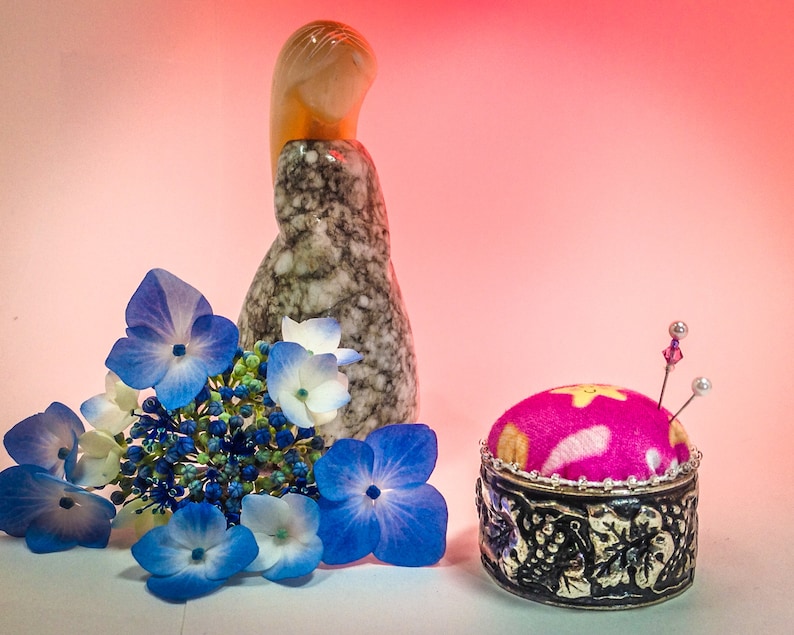 Op de afbeelding: Een stilleven met een roze en zilveren speldenkussen met spelden, een marmerpatroon beeldje en blauwe en witte hortensia bloemen. De achtergrond is een zachte roze gradi&euml;nt.