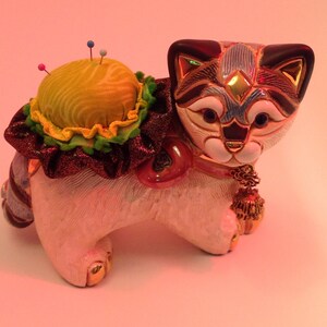 May include: A white ceramic cat figurine with black and blue markings on its face. The cat is wearing a red and green ribbon and has a yellow pincushion on its back. The pincushion has several colorful pins sticking out of it.